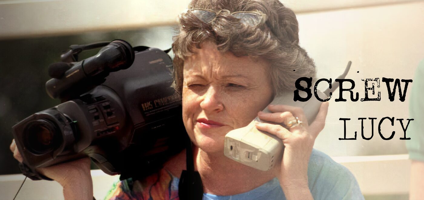 Title card for Screw Lucy: a journalist carrying a video camera on her shoulder while speaking into a mobile phone